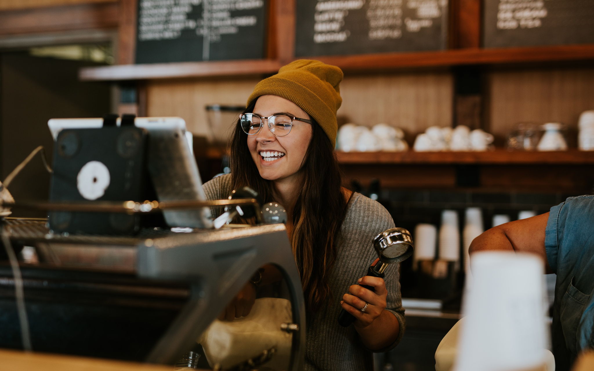 Image of a hipster girl wearing glasses and a beanie working at a coffee shop making espresso and smiling.