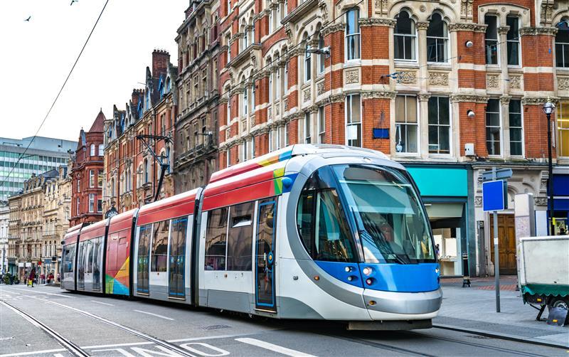 A modern light rail tram with a blue, red, and white design travels along tracks in a city center, surrounded by historic brick buildings with ornate architecture.