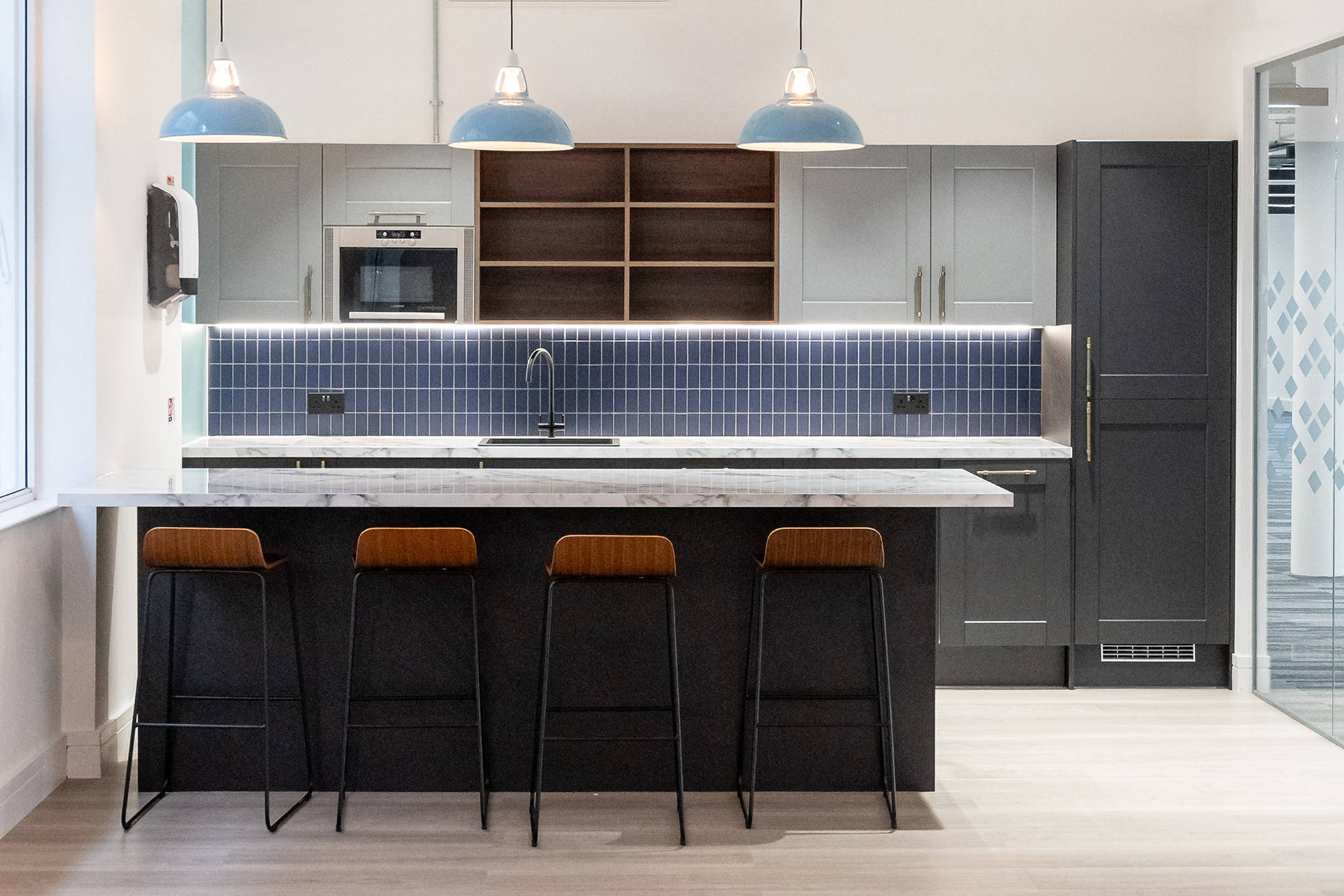 Three large Sky Blue Coolicon lampshades hang above a marble kitchen Island. With four wooden bar stools and a blue tiled back splash at the kitchen in a commercial shared office space kitchen.