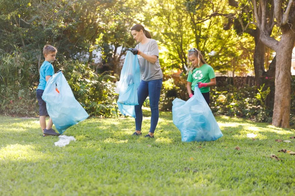 A woman and two children holding large blue trash bags while picking up litter in a park, participating in an outdoor cleanup activity on a sunny day.