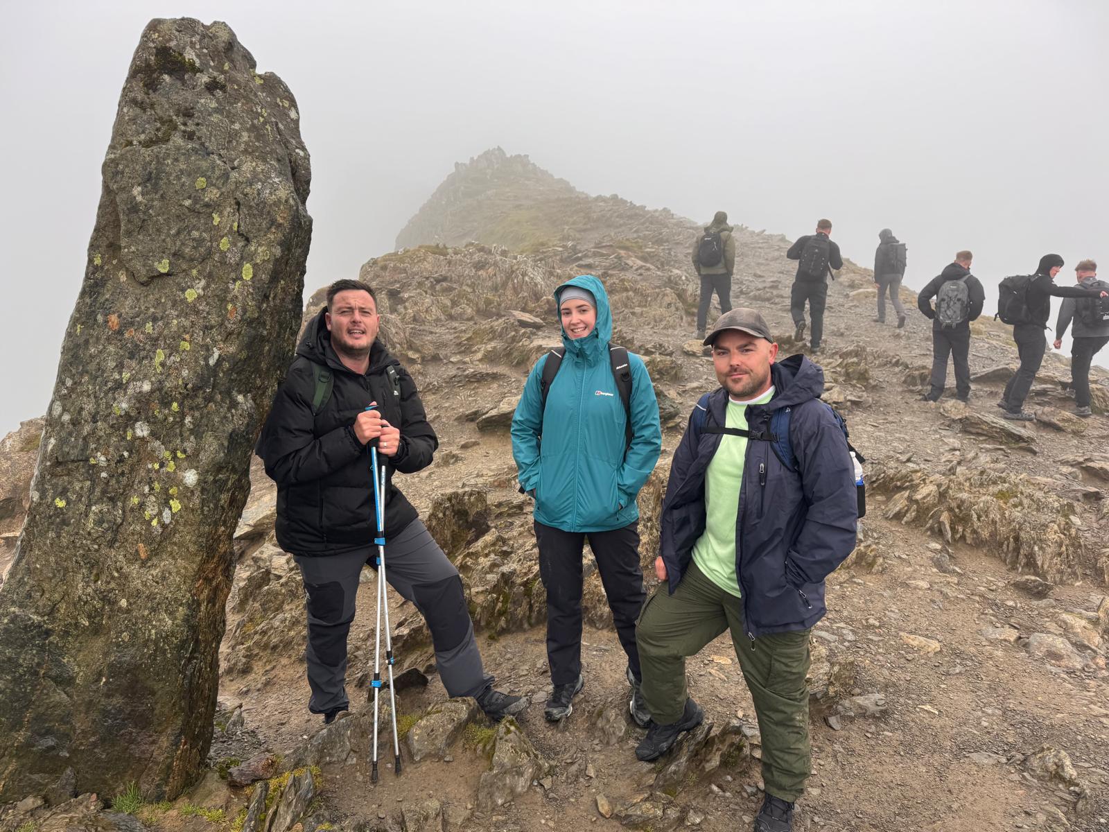 Three hikers in outdoor gear stand on a rocky, foggy mountain path next to a tall, lichen-covered stone. Several other hikers are visible in the background, walking or standing along the ridge.