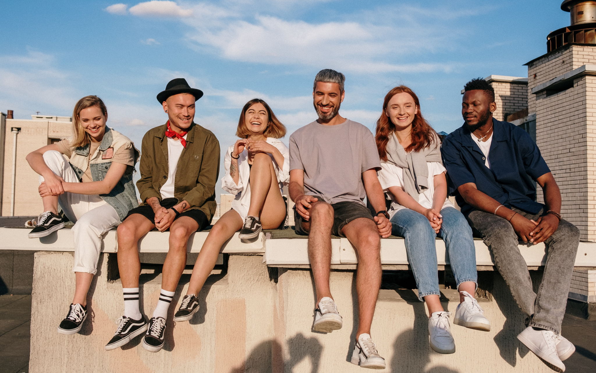 A large group of attractive and hip people smiling and sitting together on the edge at the roof of a building.