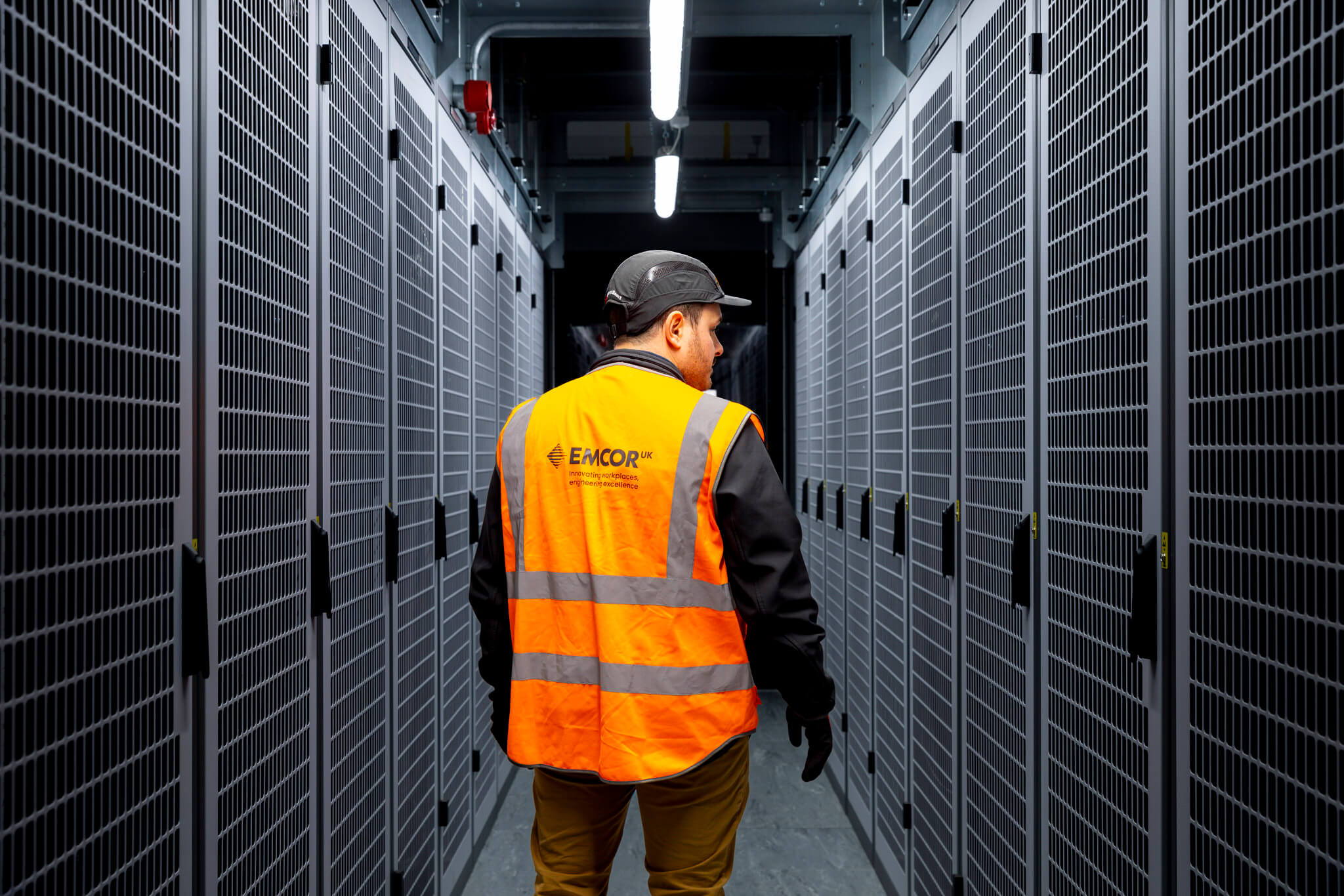 A man wearing an orange high-visibility vest and a cap walks down a corridor lined with large server racks, illuminated by overhead lighting.