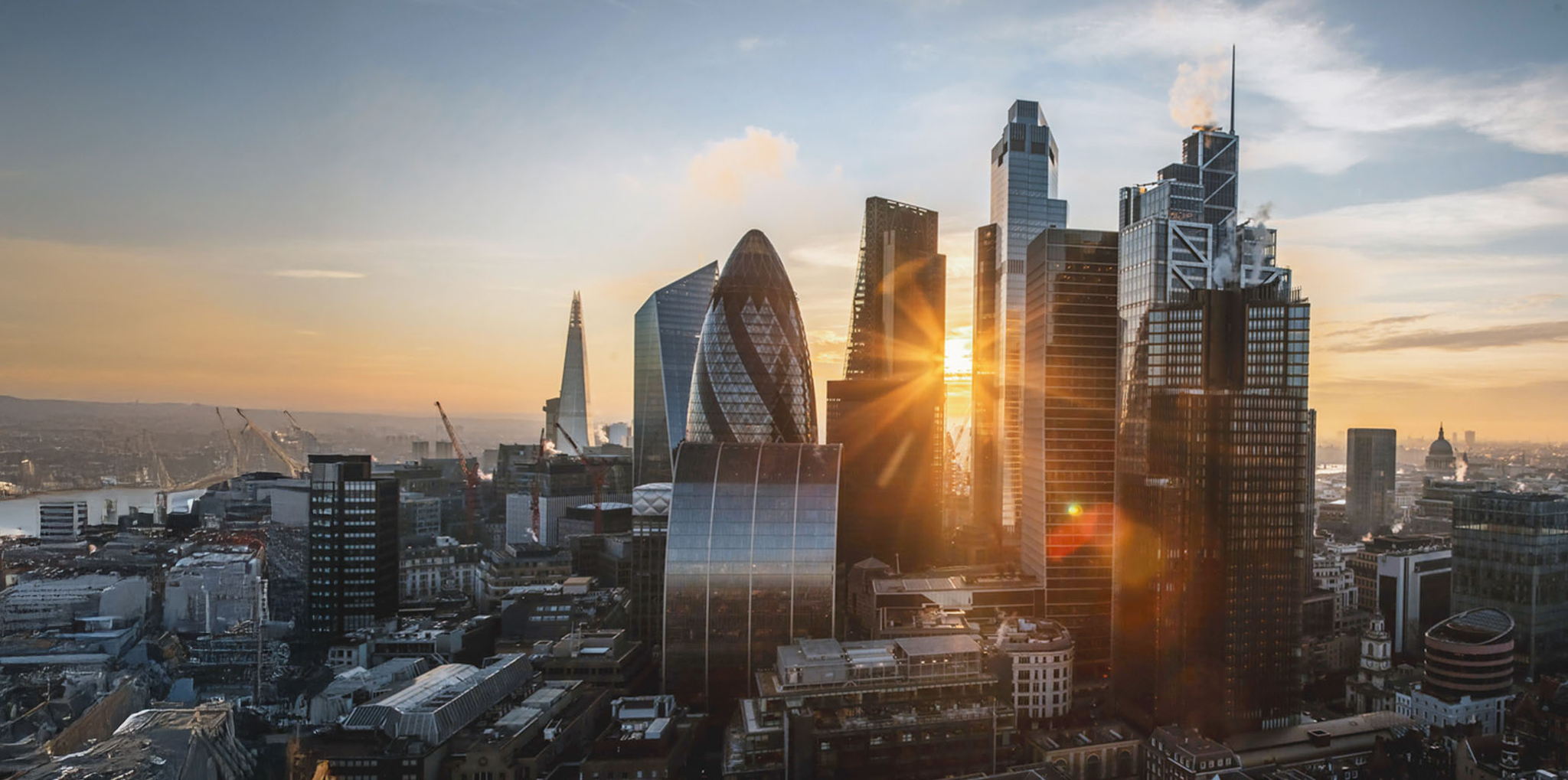 London city skyline at sunset, featuring modern skyscrapers such as The Gherkin and The Shard with sunlight reflecting off glass buildings and a warm, colorful sky in the background.