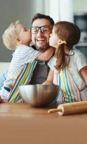 Man with glasses getting a kiss from his son and daughter while baking in the kitchen