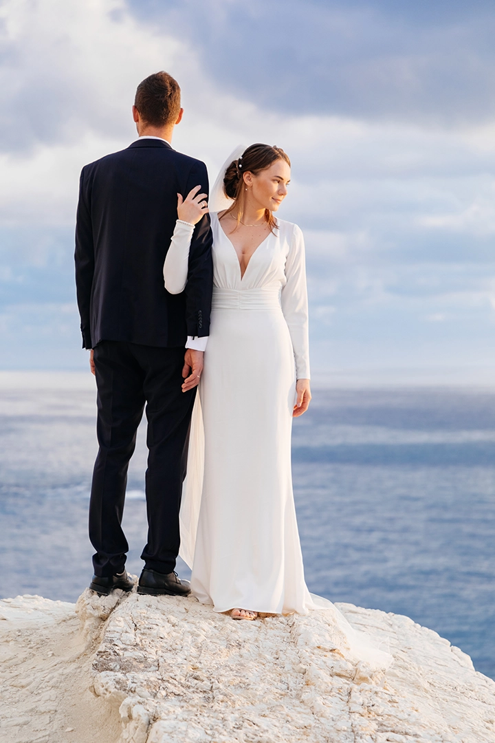 Un couple élégant vêtu de tenues de mariage modernes se tient au bord d'une falaise océanique, surplombant les vastes eaux de la mer.