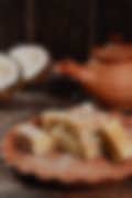 Traditional Indian coconut burfi sweets on a terracotta plate, with shredded coconut sprinkled around and a clay teapot and coconut halves in the background.