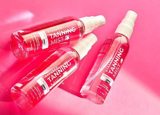 3 Flaschen Strawberry Tanning Mist–Selbstbräunungsspray vor einem einfarbigen rosa Hintergrund, der die Schnellbräunungsformel hervorhebt.