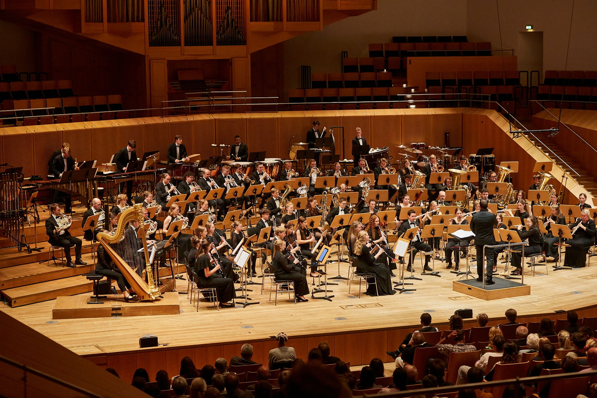 Ein großes Blasorchester musiziert in schwarzer schicker Kleidung auf einer Bühne vor Publikum. 