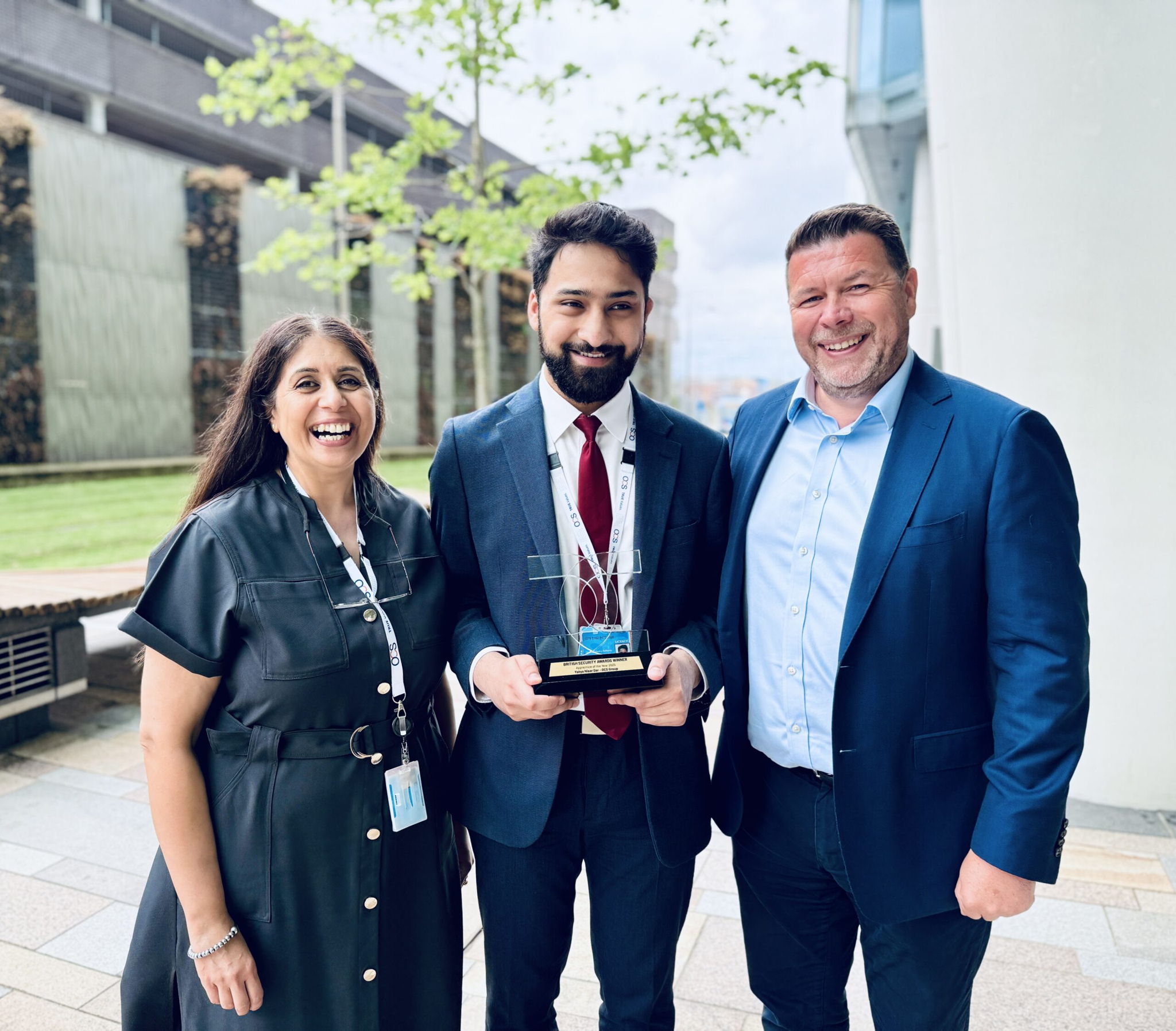 Three people are standing outside, smiling at the camera. The person in the center holds a trophy, and all are dressed in business attire. There are modern buildings and greenery in the background.