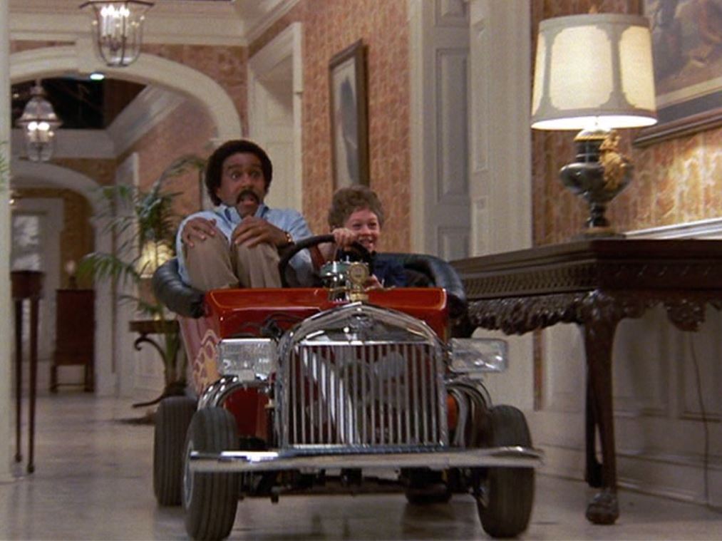Richard Pryor and Scott Schwartz driving down the hallways of a mansion in a small red toy car.