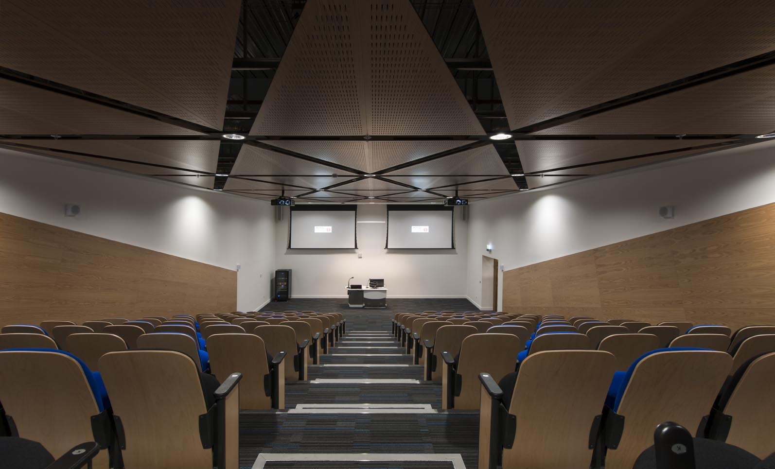 A modern, empty lecture hall with tiered seating, wooden armrests, and two large projection screens at the front above a white lectern and equipment desk. The room has angled walls and a geometric patterned ceiling.