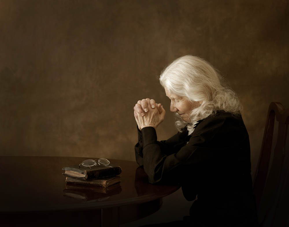 An older woman praying at her table next to the Bible.