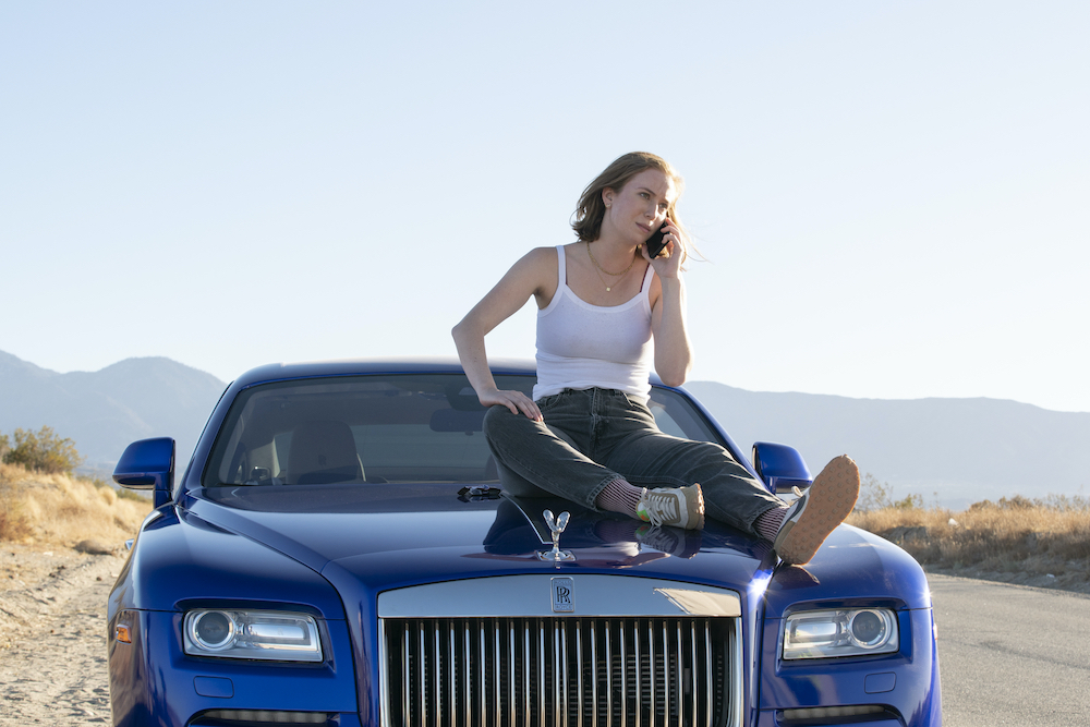Ava wearing a white tanktop and jeans, sitting on top of sleek blue car, and is on the phone with a concerned look.