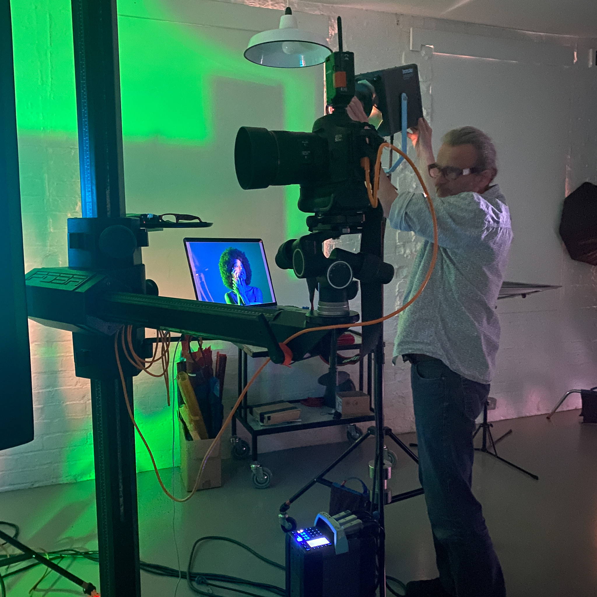 Photographer Carl wears a white shirt with blue jeans and thick black glasses, he is adjusting a piece of lighting equipment. In the foreground, a camera is mounted on a large gimbal that stands to the left. In the background, a laptop rests on a trolley a shows the close-up head-shot of a model bathed in green and blue lighting. 