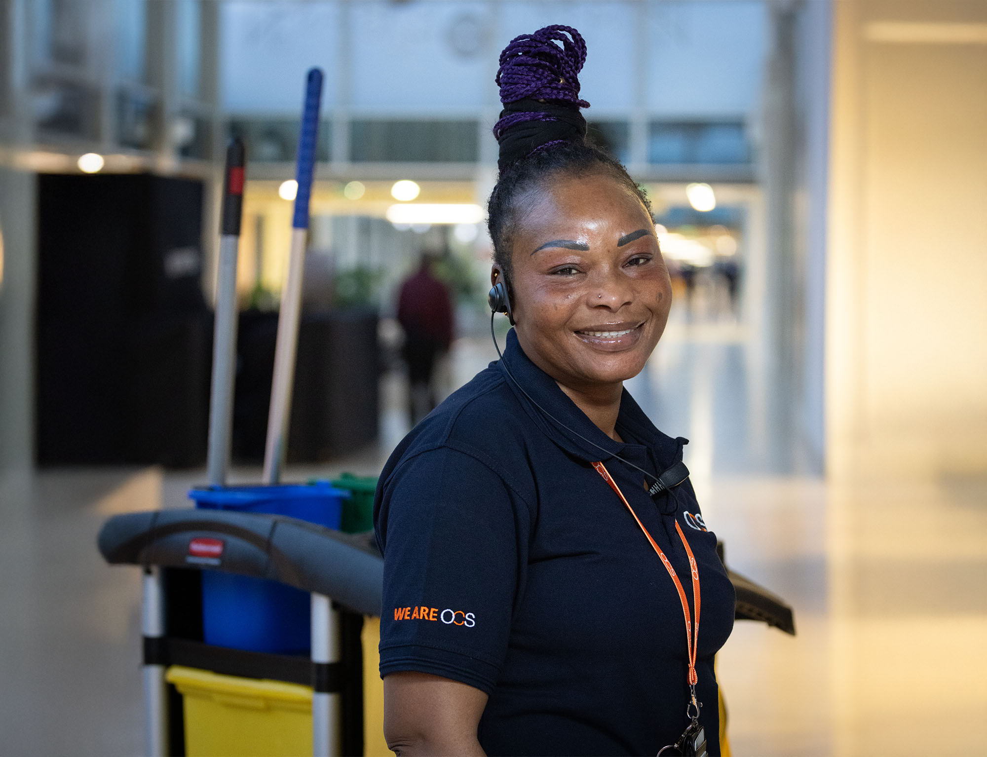 A smiling woman wearing a dark polo shirt and headset stands in a hallway next to a cleaning cart with mops and yellow and blue buckets. Her hair is styled in braids tied up in a bun.