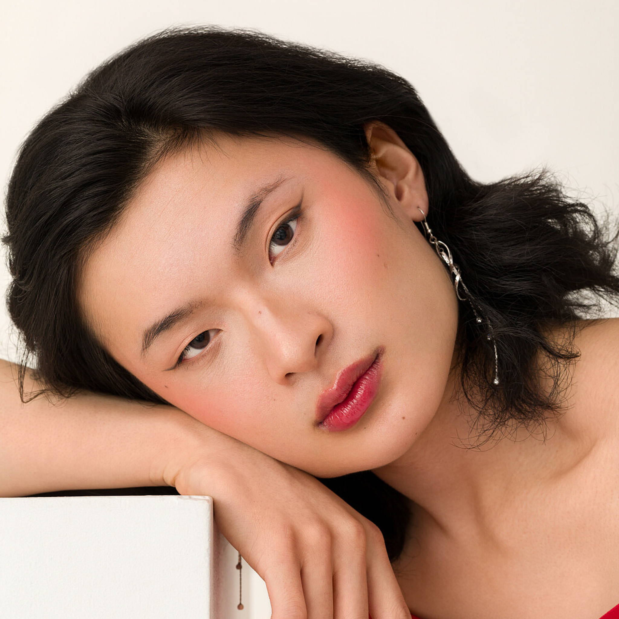 Model Jaden is facing the camera against a white background. Their head is propped up on the back of their right hand which is resting on a white box. They are wearing red lipstick and a long, delicate metal earring.