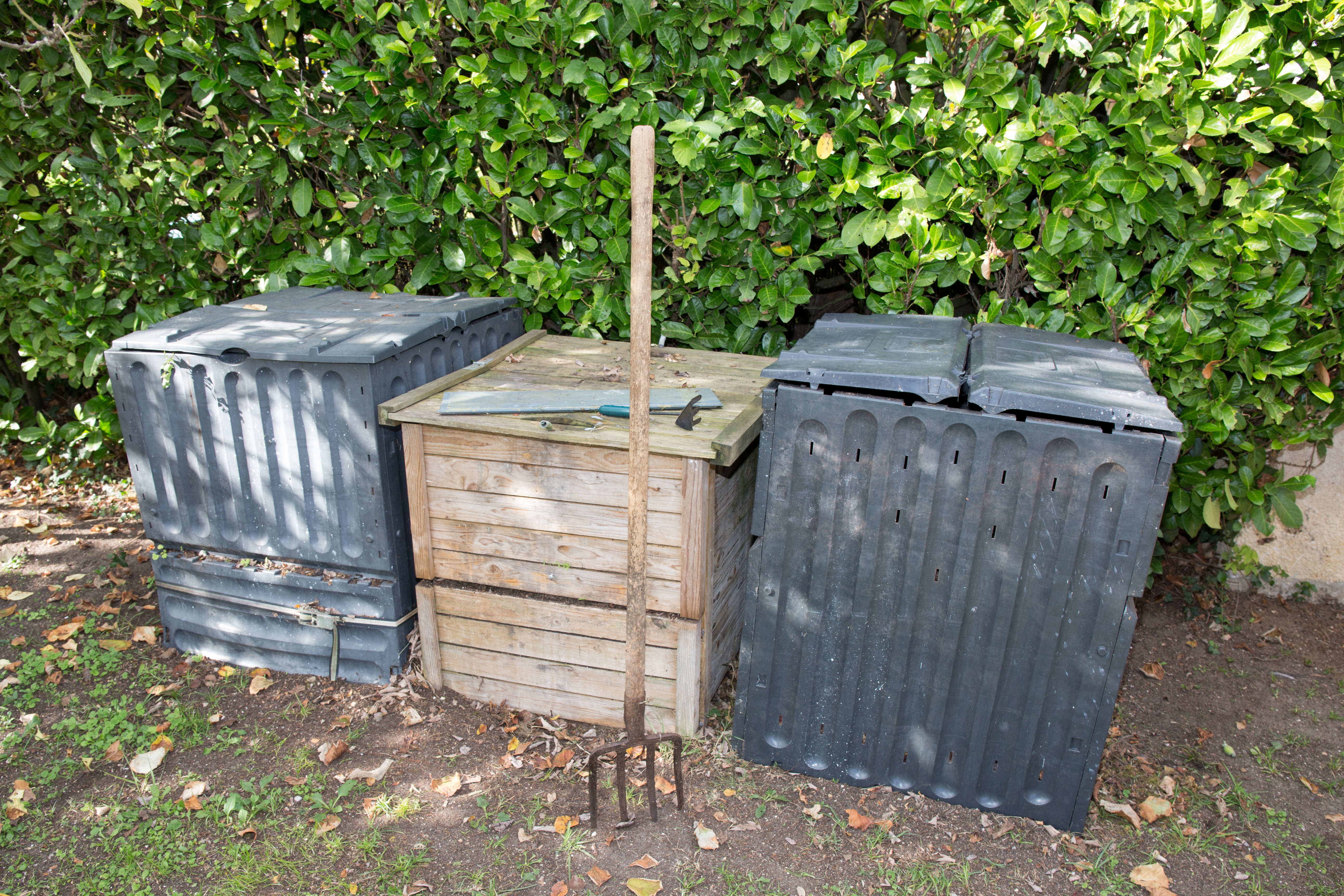 Three compost bins made with different materials side-by-side with a garden fork in the foreground and a hedge in the background