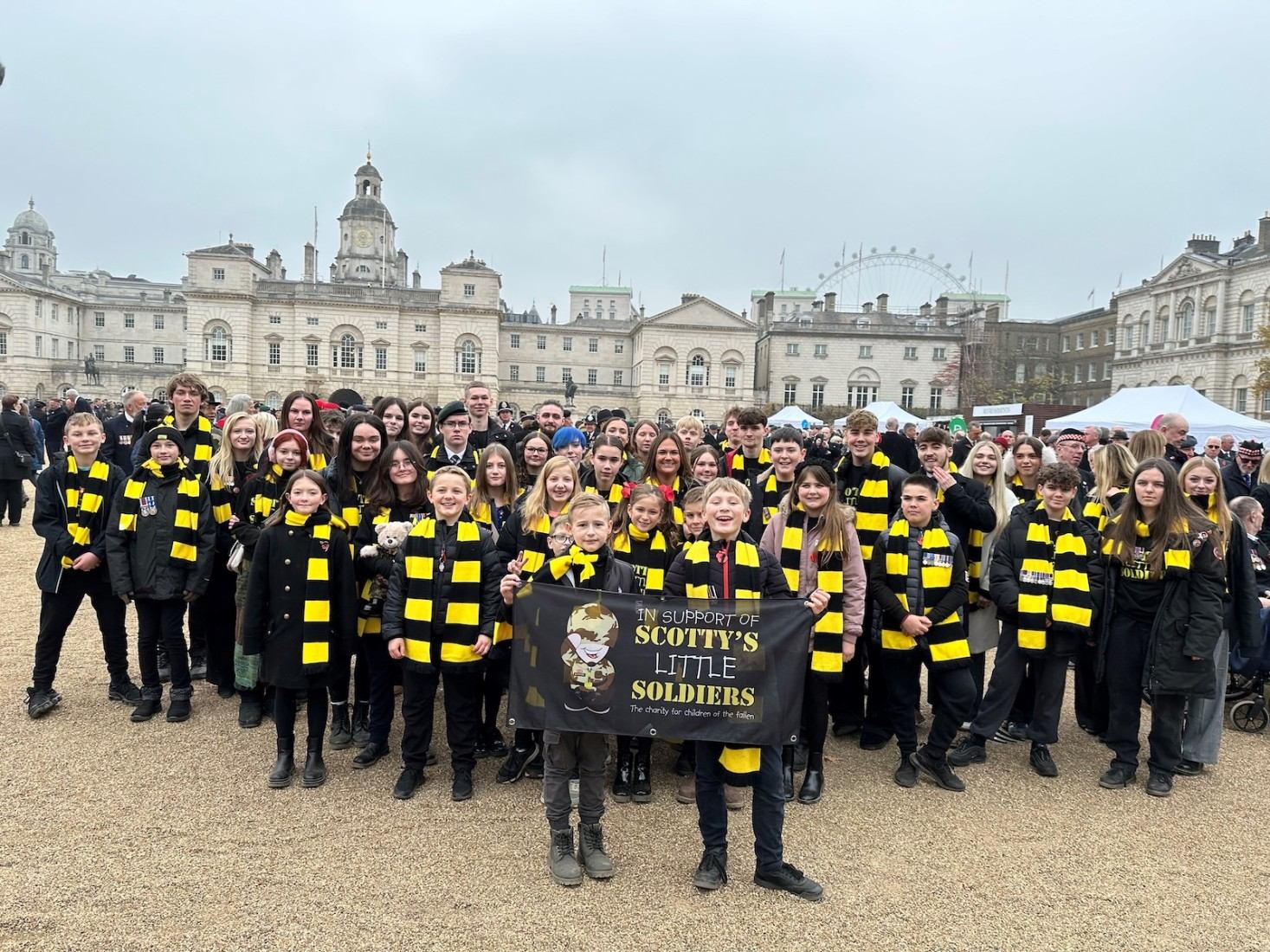 A large group of children and teens wearing yellow and black striped scarves stand together outdoors, holding a banner that reads “In Support of Scottys Little Soldiers.” Historic buildings are visible in the background.