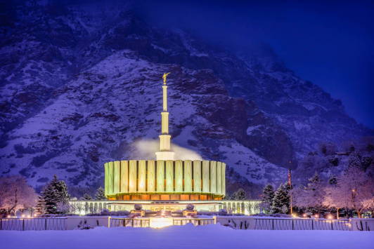 Provo Temple glowing against the wnoy mountains at night.