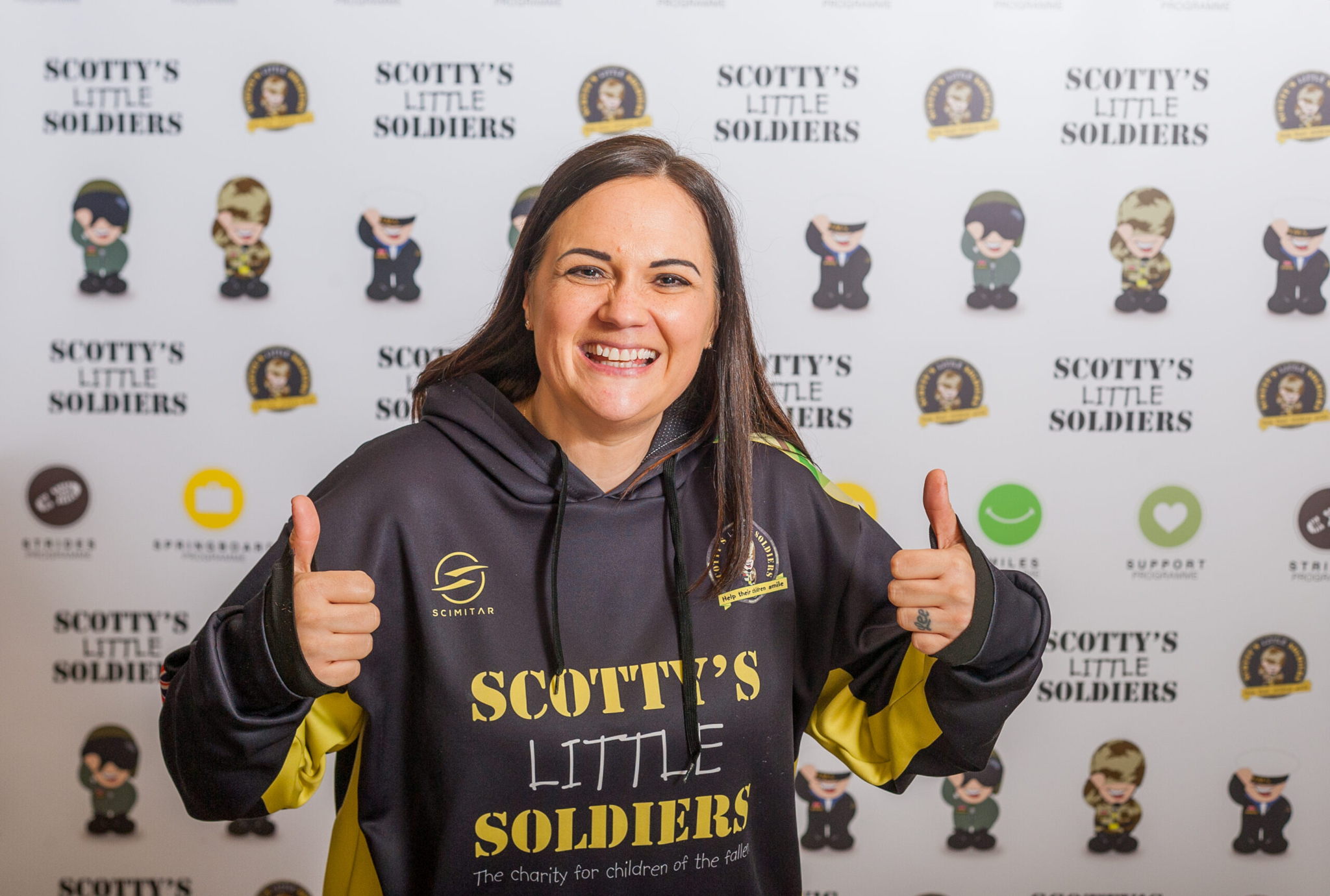 A smiling woman gives two thumbs up, wearing a Scottys Little Soldiers hoodie, standing in front of a branded backdrop with logos and cartoon soldiers.