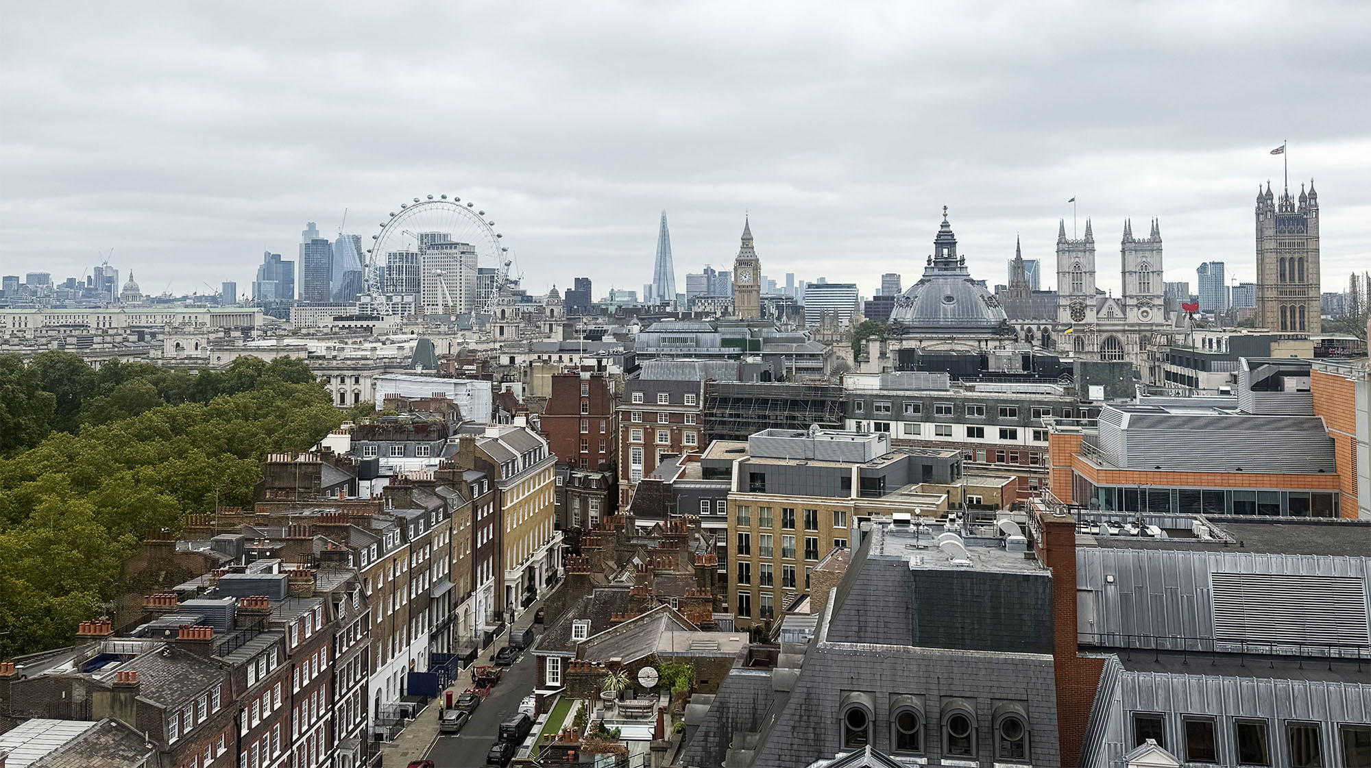 A cityscape of London featuring the London Eye, the Shard, and Westminster Abbey under a cloudy sky, with a mix of historic and modern buildings visible in the foreground.
