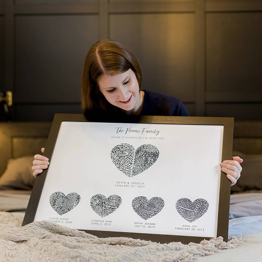 A couple embraces affectionately in front of a framed artwork featuring two overlapping hearts signed by guests. The mood is loving and intimate.