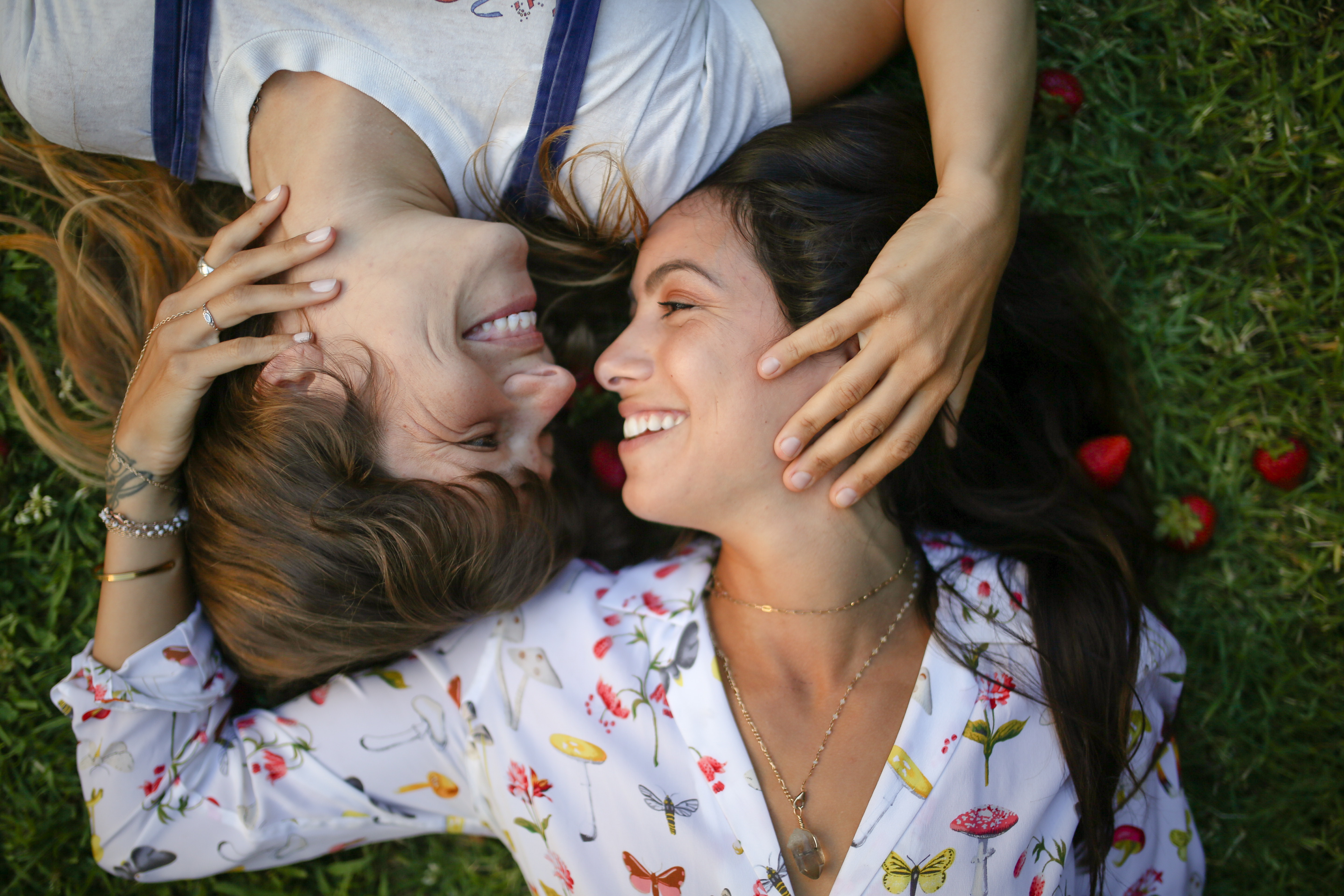 Two attractive women wearing stylish outfits lying on the grass together with one hand on eachothers' faces.