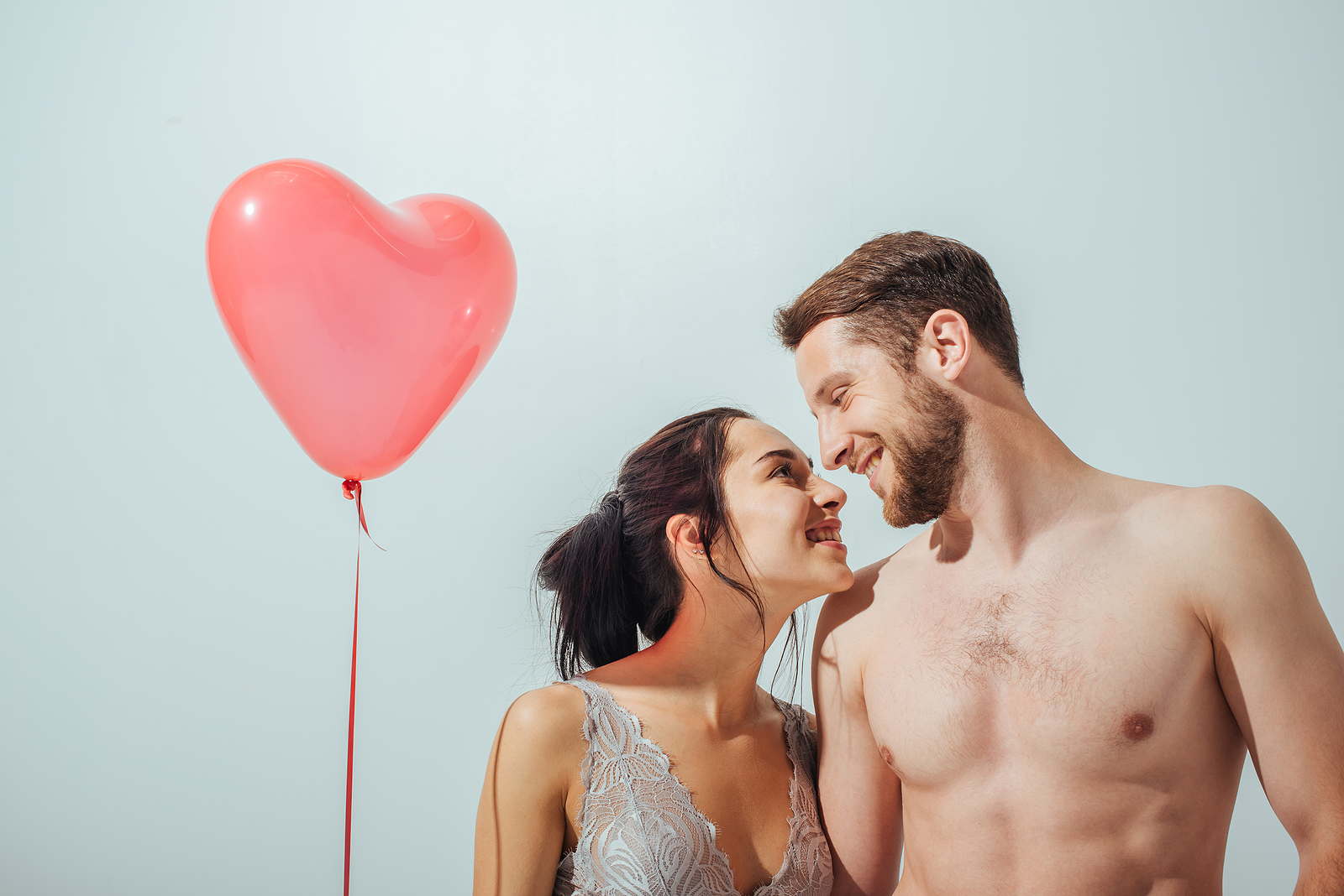An attractive couple stands close together wearing underwear looking into eachother smiling holding a heart shaped balloon.