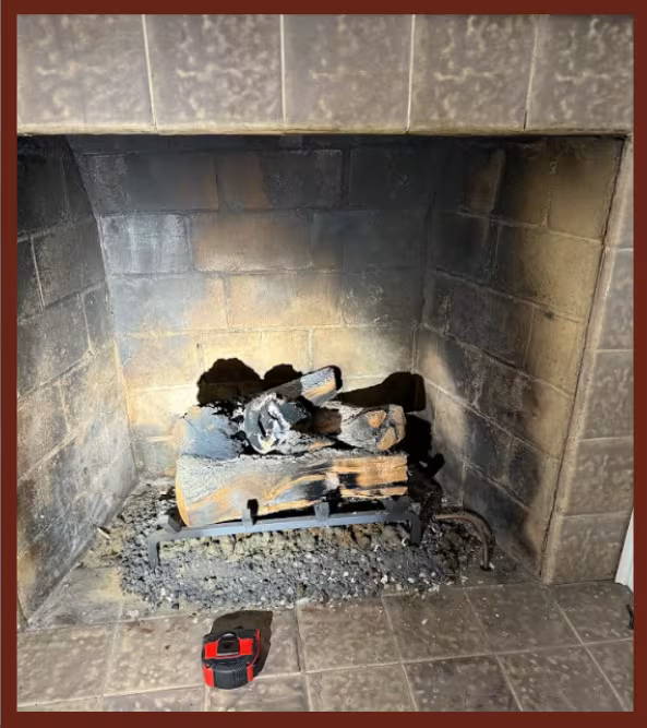 Soot-stained fireplace firebox with damaged bricks and charred logs on a metal grate, showing signs of wear and needed repair.