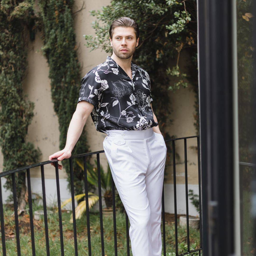 man wearing white pants and a gothic floral short sleeve silk shirt while standing on a terrace