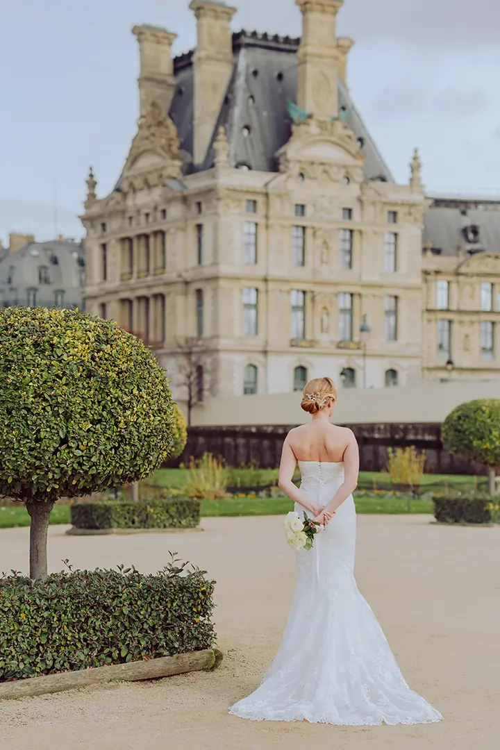 La mariée debout de dos face au bâtiment du musée du Louvre à Paris. Elle tient un bouquet de fleurs et arbore une coiffure relevée.