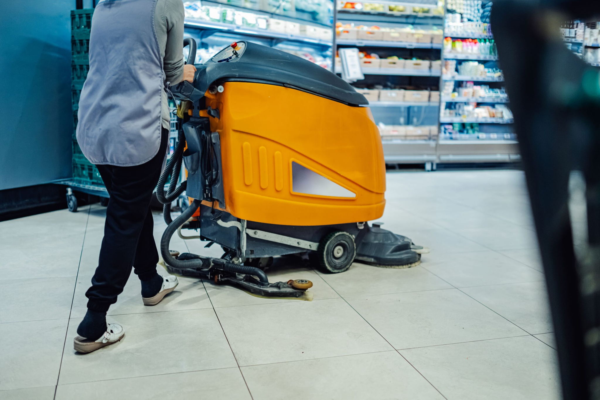 A person operates an orange floor cleaning machine in a grocery store aisle, cleaning the tiled floor near shelves stocked with products.