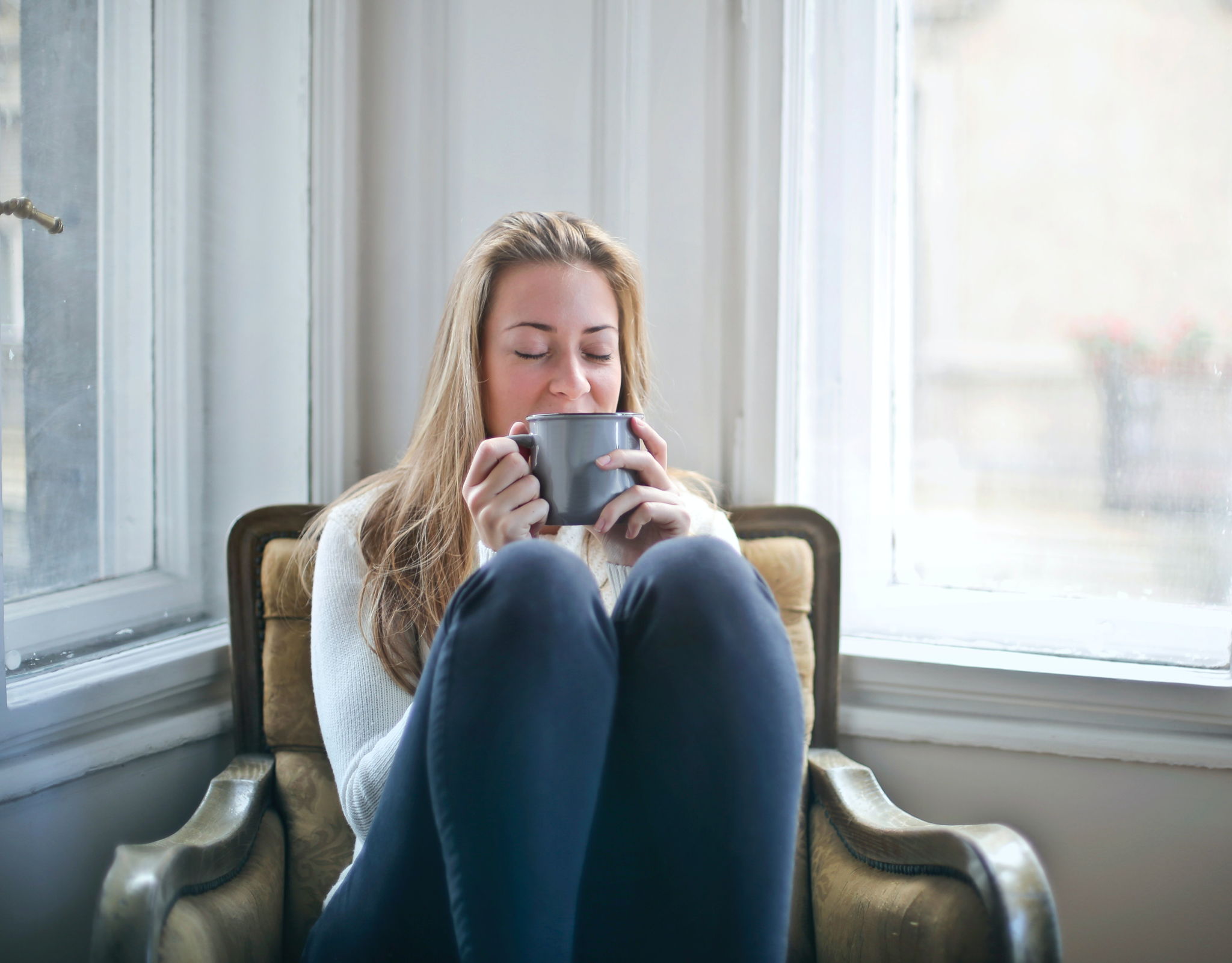 A blond haired woman is sitting with her knees up on a chair with a coffee mug close to her nose.