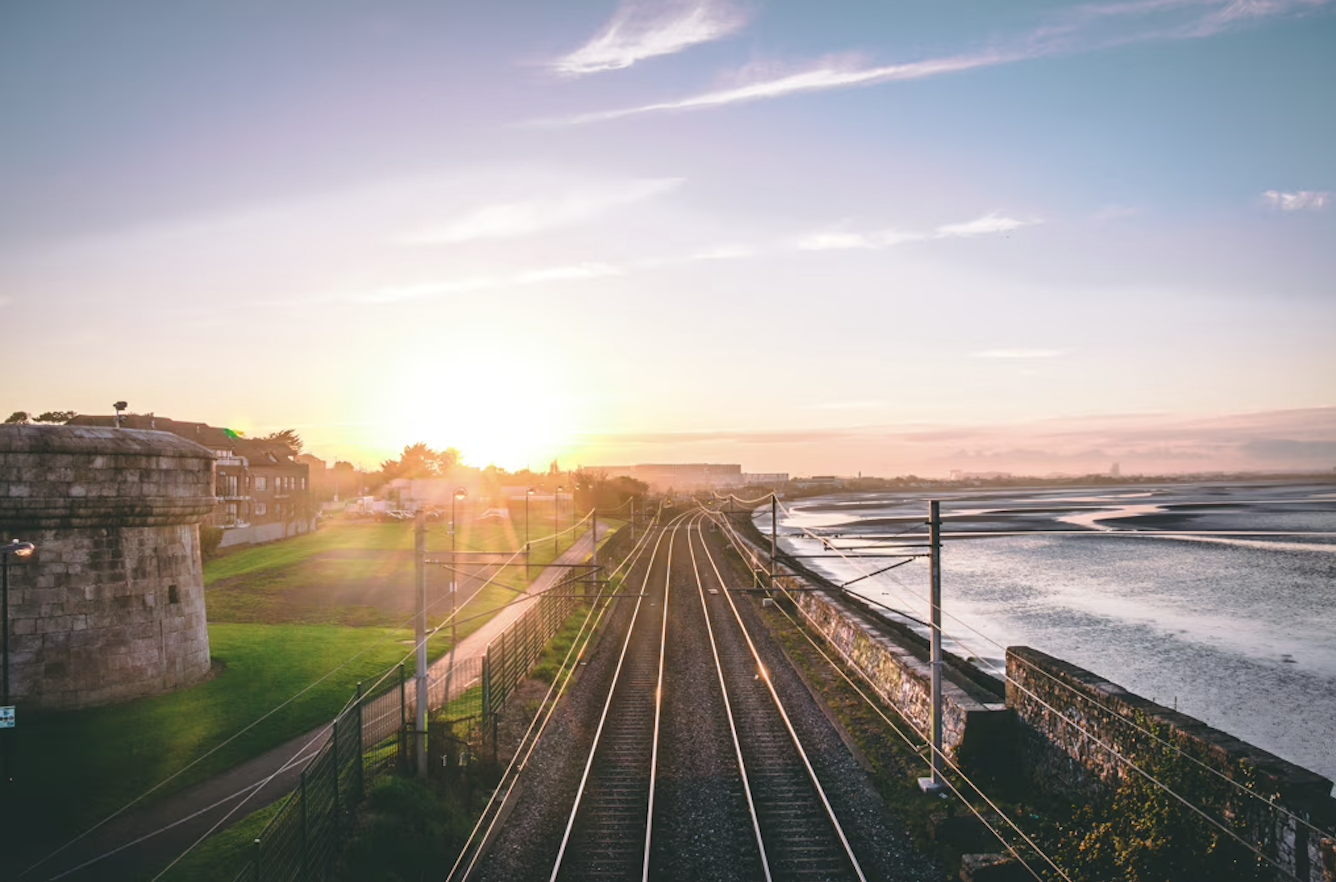 Railway tracks run parallel toward the horizon at sunset, with the sun low in the sky. To the left are buildings and a round stone tower; to the right is a coastline with calm water. The scene is serene and bathed in warm light.
