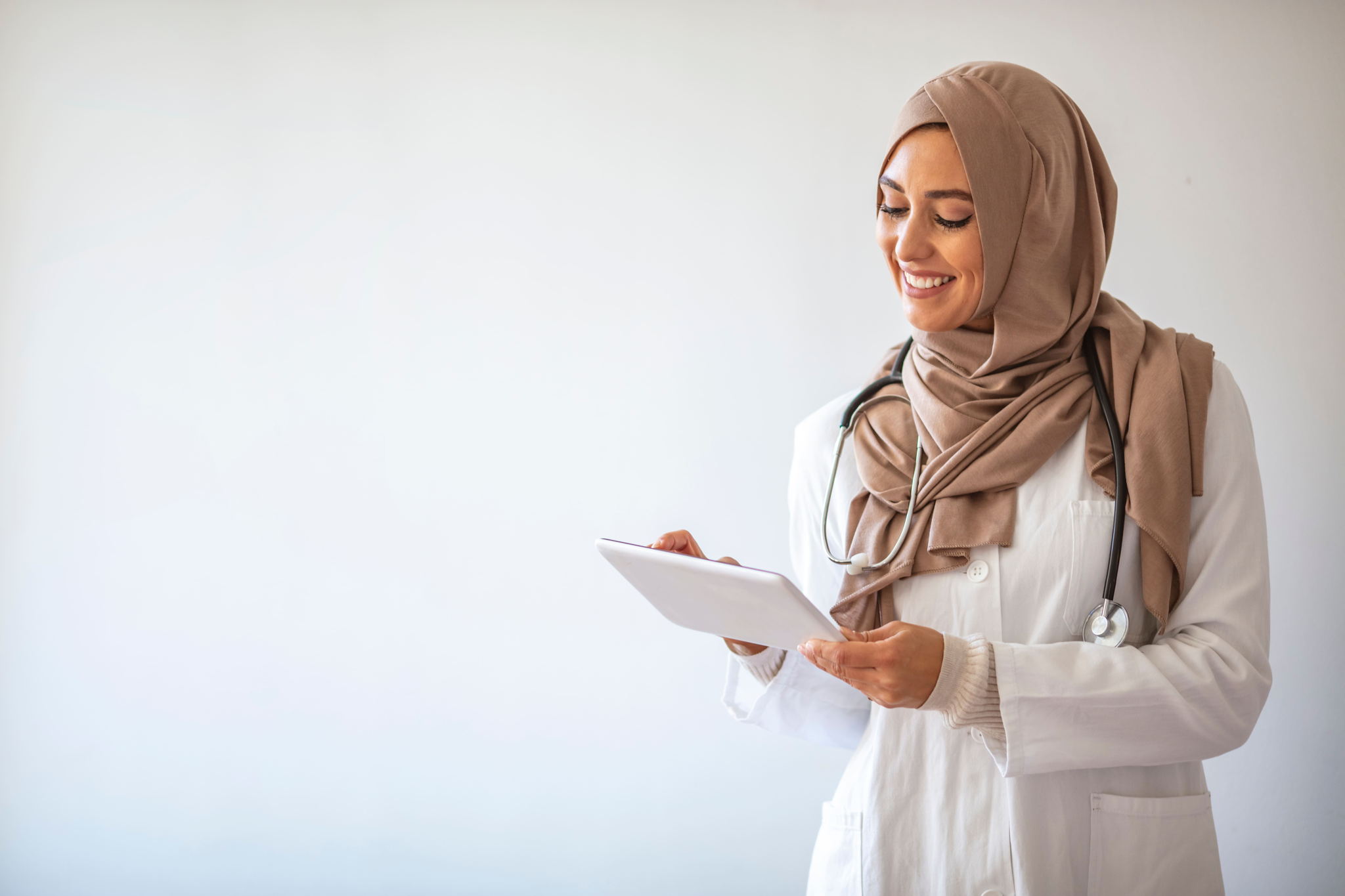 A smiling female doctor wearing a tan hijab and white lab coat holds a tablet, looking at the screen. She has a stethoscope around her neck and stands against a plain, light background.