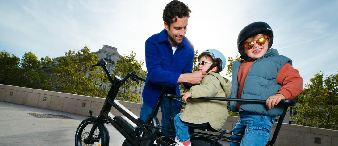 Een vader zet zijn kinderen achterop zijn elektrische bakfiets.