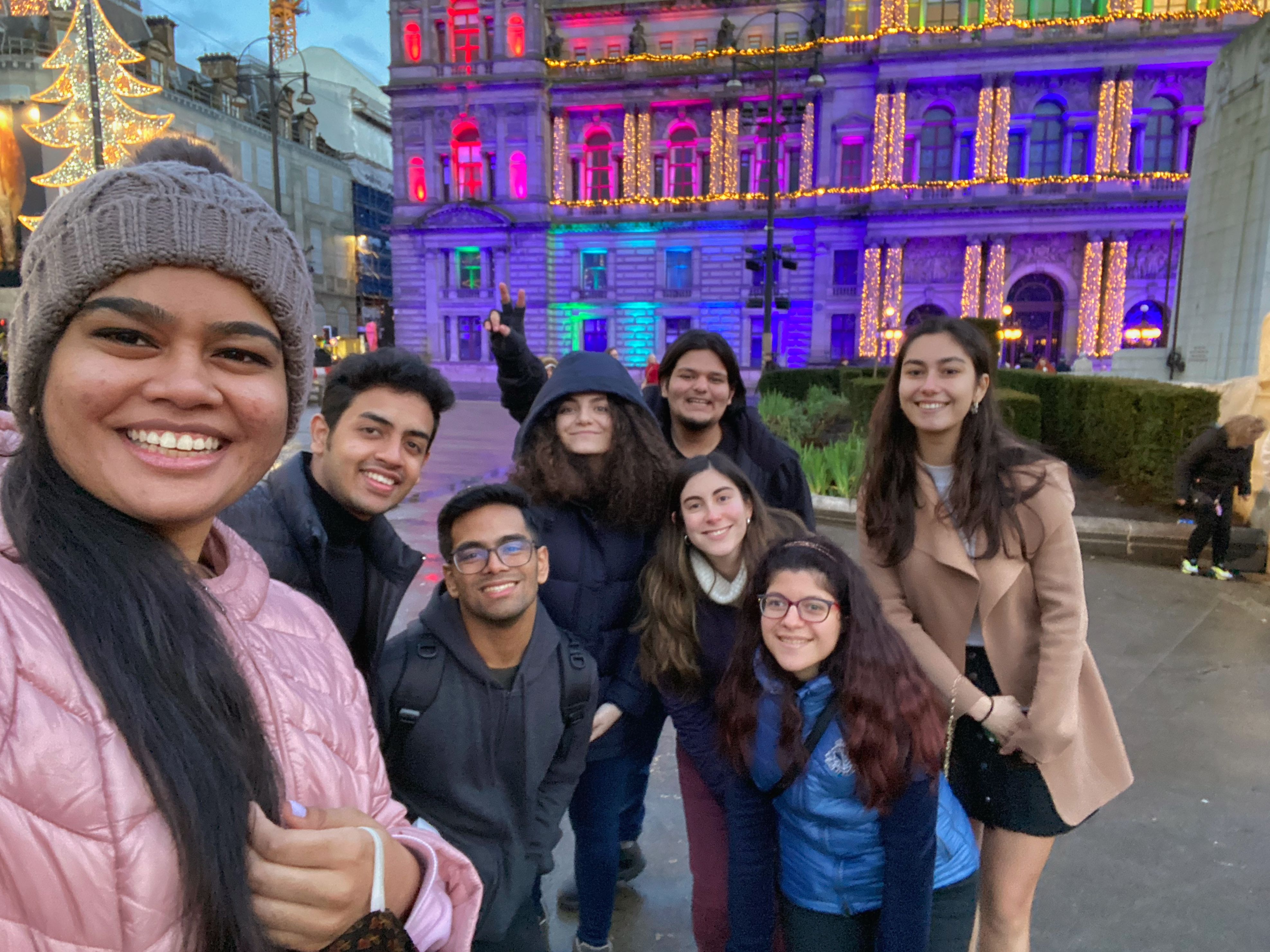 A group of friends stand together smiling in winter clothes. Rishabh is at the back making a peace sign. There is a building in the background which is lit in urple, blue, pink and yello and it looks festive.