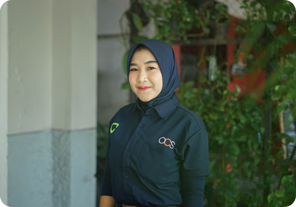 A person wearing a blue hijab and a blue shirt stands smiling. The shirt features green and orange logos. The background includes greenery and a white wall.