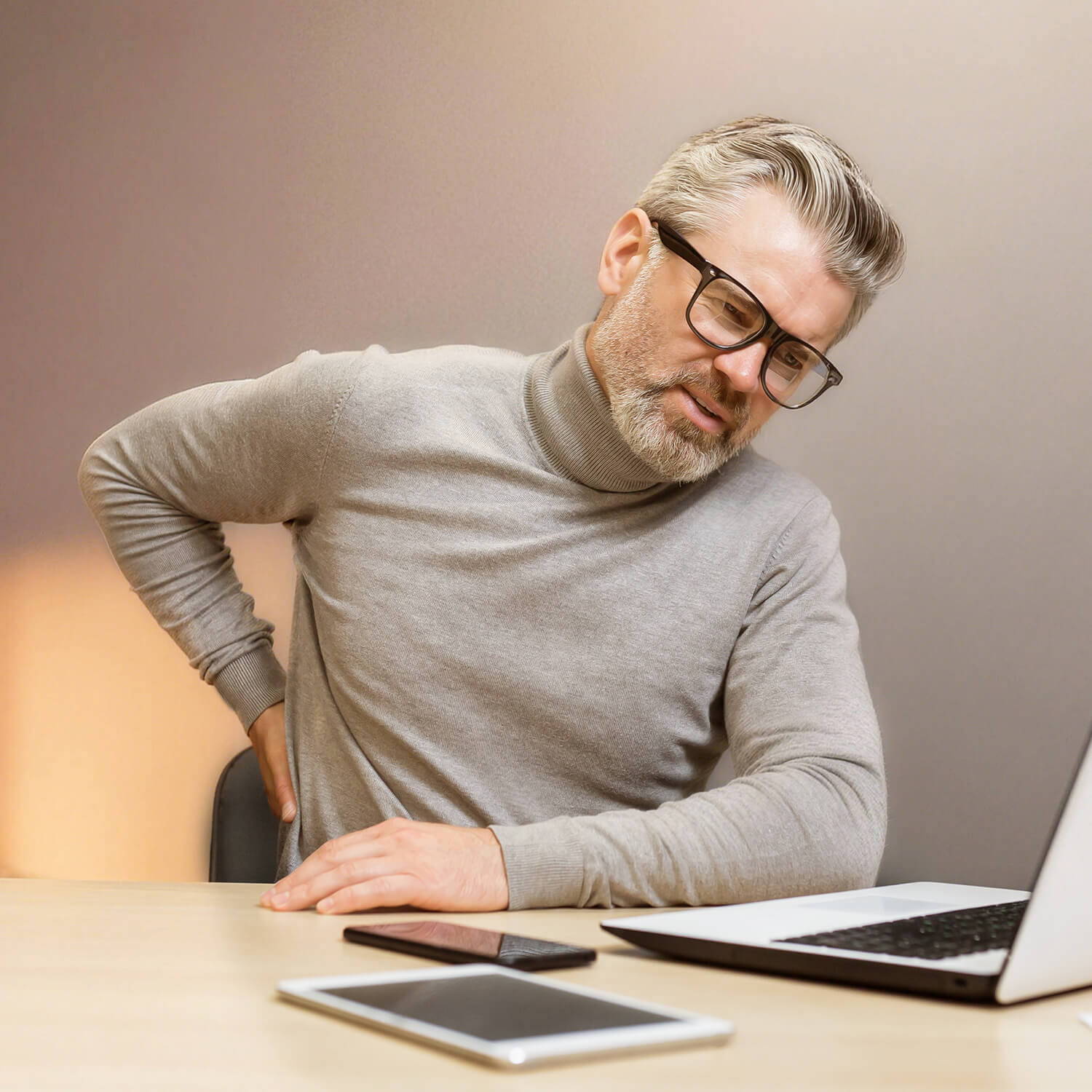 Middle-aged man sitting at a desk holding his lower back, showing everyday back pain from long hours at work