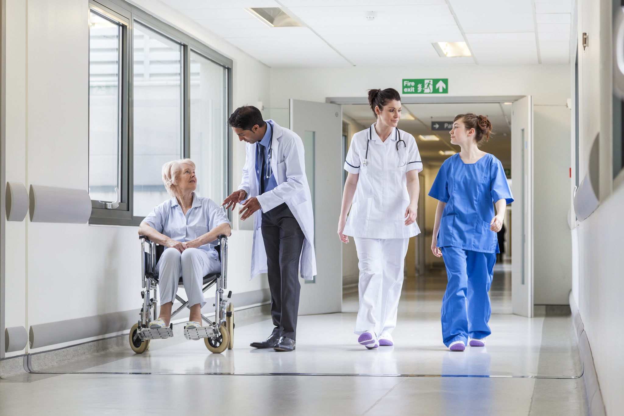 A doctor speaks with an elderly woman in a wheelchair while two nurses walk alongside them in a bright hospital hallway.
