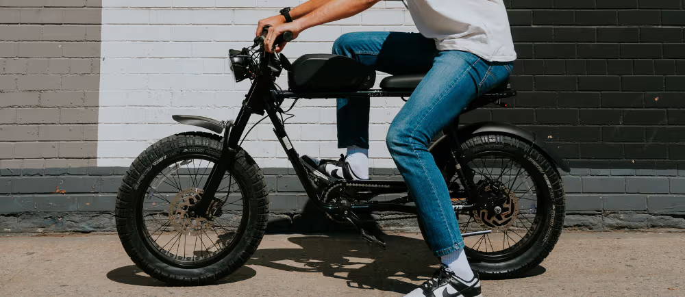 a side view of a man in jeans and a white t shirt sitting on an e-moto in front of a white wall