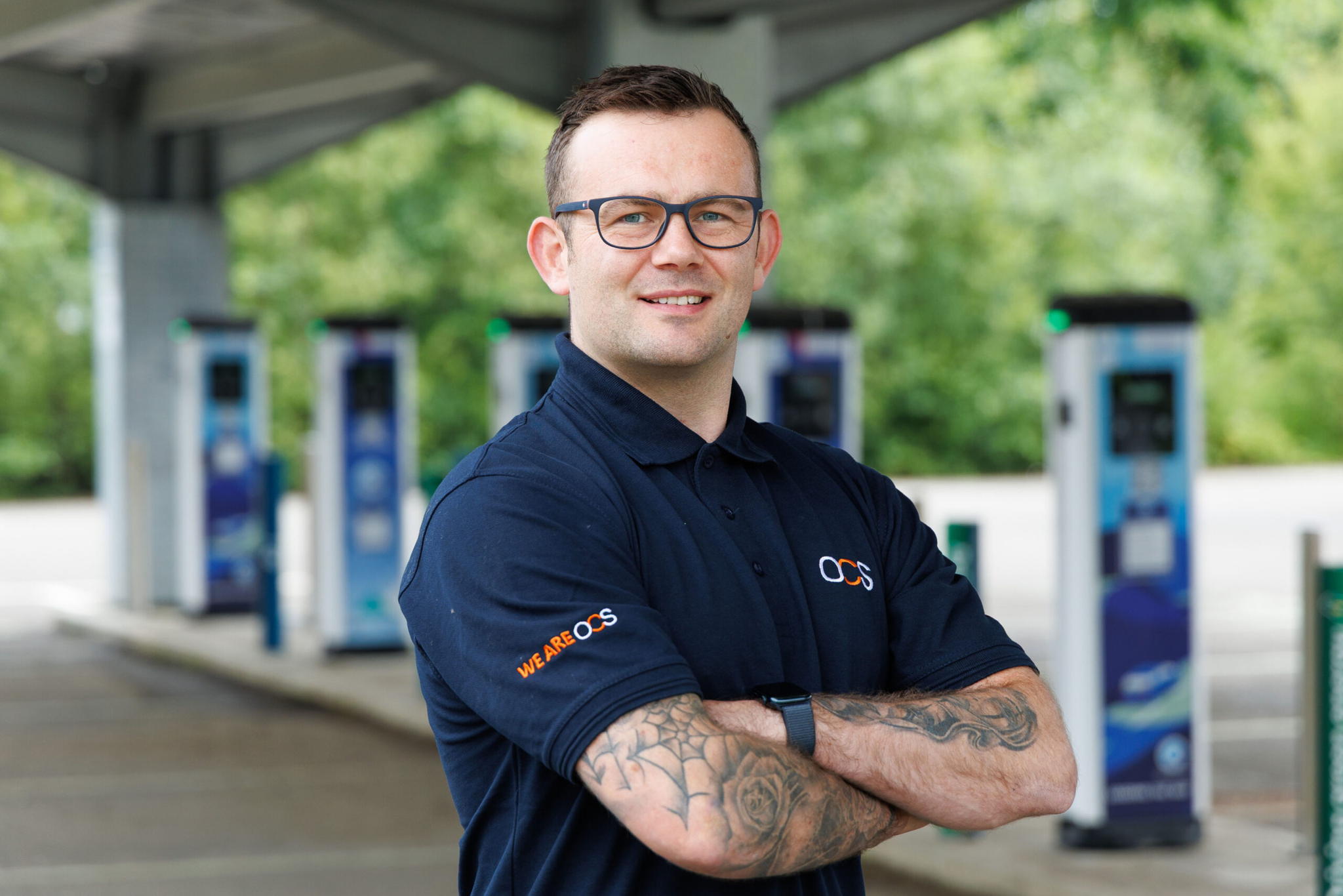 A man with glasses and tattoos, wearing a navy blue polo shirt, stands with arms crossed and smiles at the camera in front of electric vehicle charging stations outdoors.