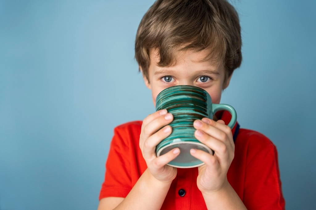 Jeune garçon en chemise rouge buvant dans une grande tasse en céramique rayée, devant un arrière-plan bleu.
