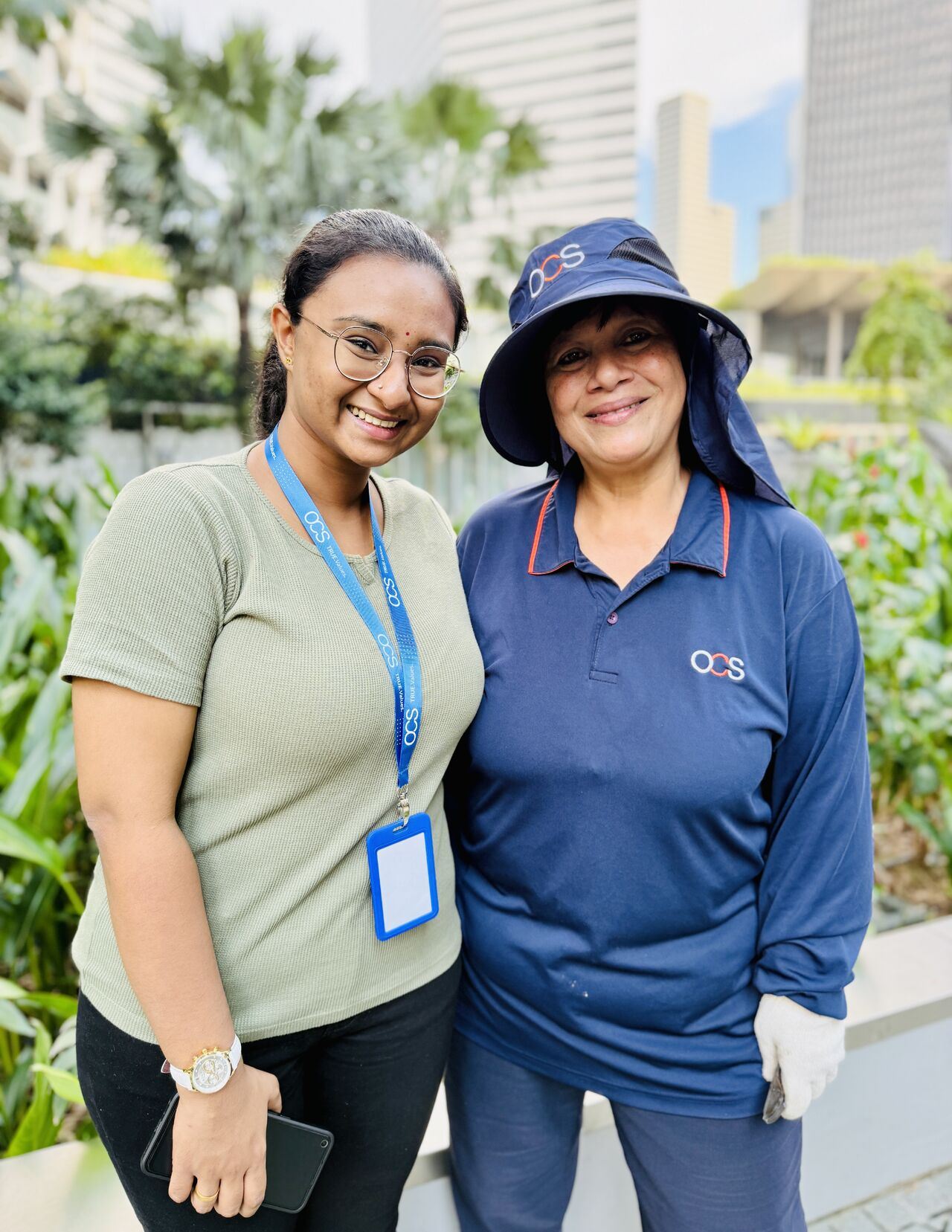 Two women smiling outdoors; one wears a green shirt and glasses with an ID badge, the other is in a blue OCS uniform with a sun hat and gloves. Green plants and city buildings are visible in the background.