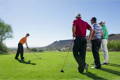 Group of friends standing together watching a golfer take a tee shot on the fairway.