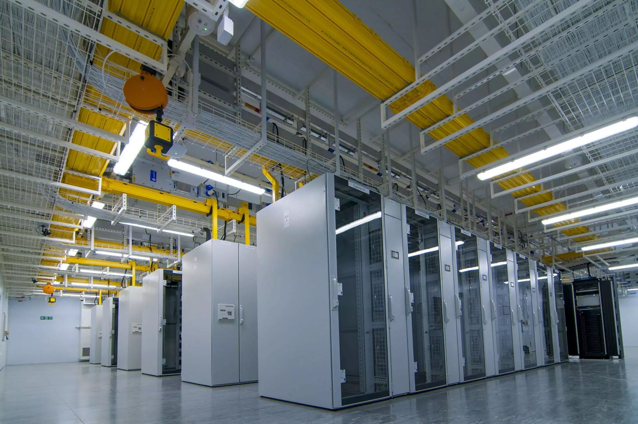 A modern data center with rows of white server cabinets, overhead yellow cable trays, and bright ceiling lights, creating a clean and organized high-tech environment.