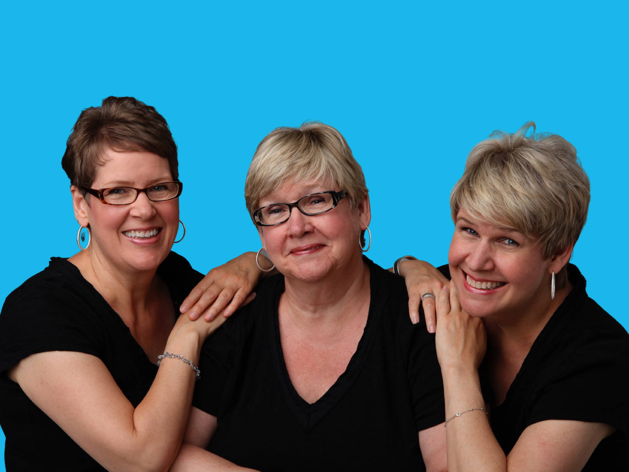 3 blonde sisters smiling in black t-shirts. the two younger sisters are leaning on the shoulders of the older sister who is seated in the middle. Image set against a blue backdrop.
