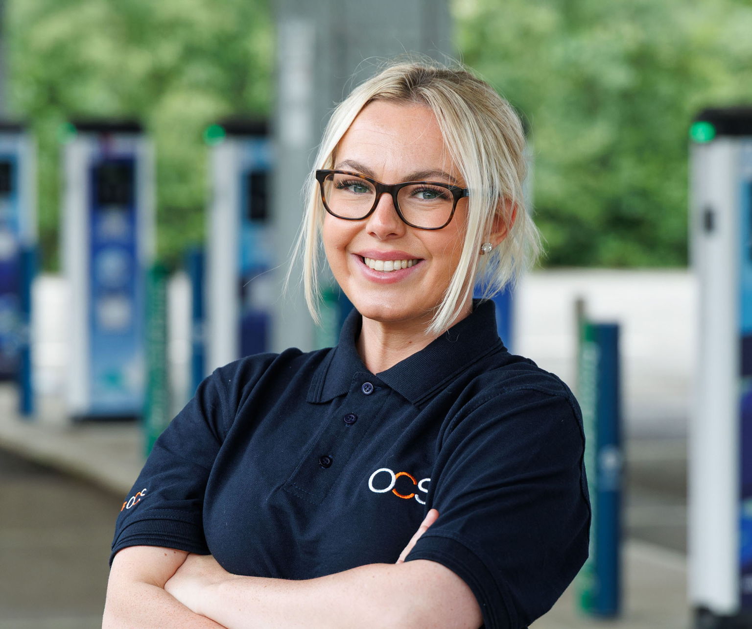 A woman with blonde hair and glasses, wearing a navy blue OCS polo shirt, stands smiling with her arms crossed at a location with several electric vehicle charging stations in the background.
