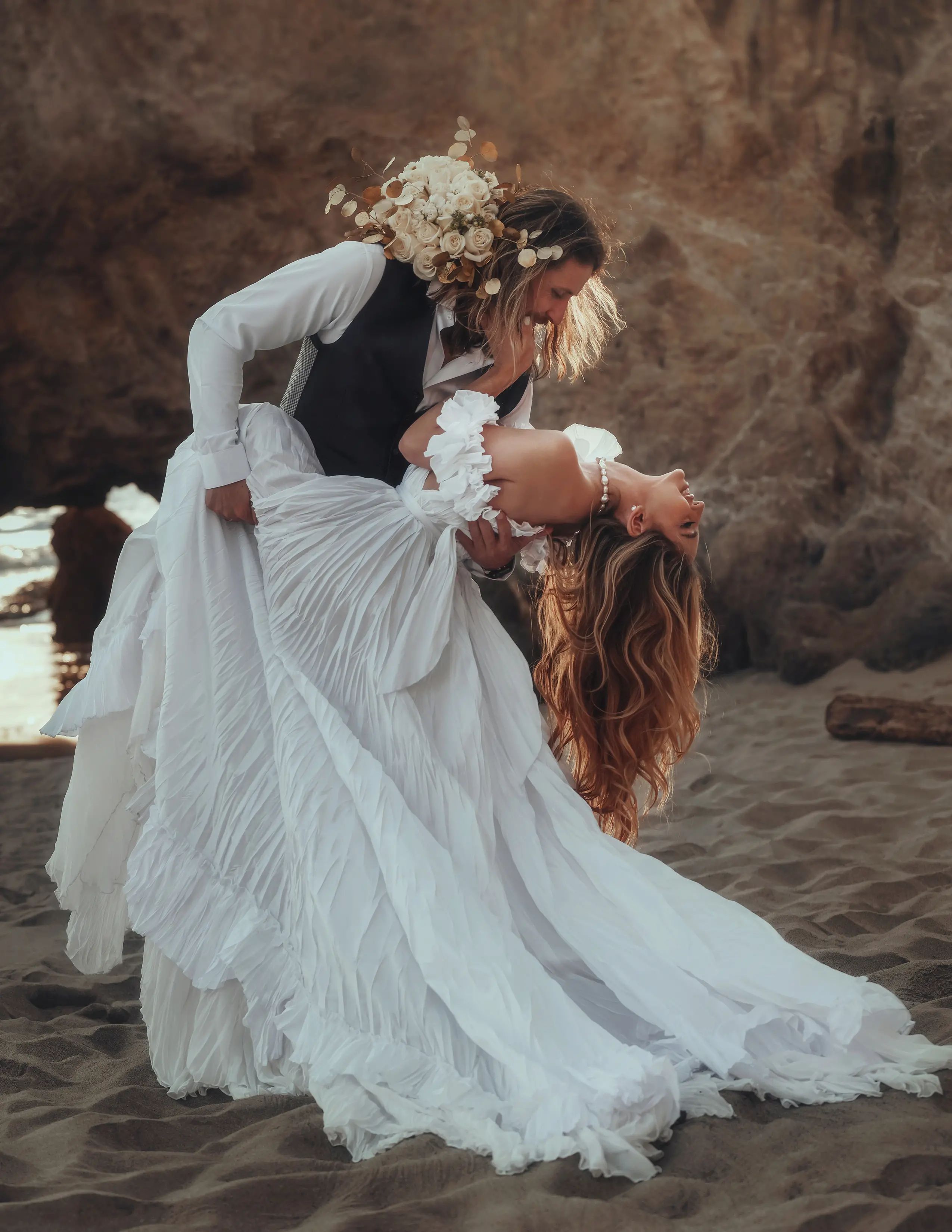 Une belle mariée aux longs cheveux portant une robe de mariée bohème blanche qui retombe sur le sable de la plage de Californie, tandis qu'elle est soutenue par le marié qui a ses bras autour de ses épaules.